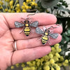 A pair of bee-shaped earrings held in a person's hand, with blurred green foliage and yellow flowers in the background.