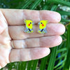 Yellow bird-shaped earrings held between fingers with a green leafy background