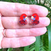 Red parrot-shaped earrings held between fingers with a green leafy background