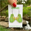A hand with red nail polish holds a white card displaying a pair of green frog-shaped earrings with orange patterned details, set against an outdoor background of green foliage and a stream.