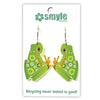 A pair of green frog-shaped dangling earrings with white polka dots and orange webbed feet, displayed on a white card with the Smyle logo and the phrase 'Recycling never looked so good!'.