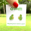 A hand with red nail polish holds a white display card featuring two small green frog-shaped stud earrings. The card shows the brand name 'smyle designs' and the slogan 'Recycling never looked so good!'.