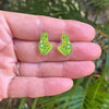 Green frog-shaped earrings held between fingers with a natural background