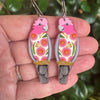 A pair of handcrafted parrot earrings held in a person's hand, featuring pink and orange parrot bodies with colorful floral patterns and gray beaks and tails, set against a blurred green background.