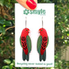 Red and green bird-shaped earrings on a Smyle Designs card with a green leafy background.