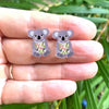 Two koala-shaped earrings held between fingers with a natural background