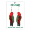 A pair of handmade parrot-shaped earrings on a white backing card. The earrings are made from recycled materials, featuring a red top, green wings, and black patterned details.