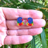 A close-up of a hand holding a pair of colorful parrot stud earrings against a blurred green outdoor background.