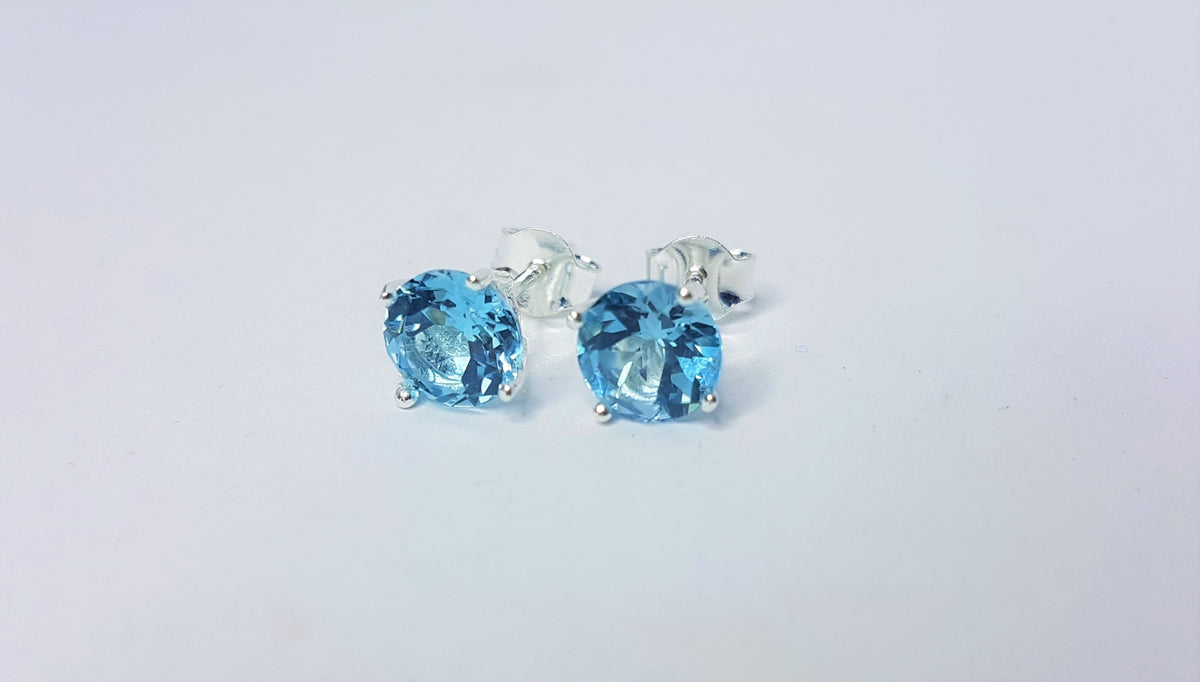 Pair of 7mm round blue topaz claw-set sterling silver stud earrings on a light gray background