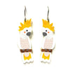 A pair of dangling earrings shaped like cartoon cockatoos, with yellow spiky feathers, white bodies, pink patterned clothing, and brown perched details, shown on a plain white background.
